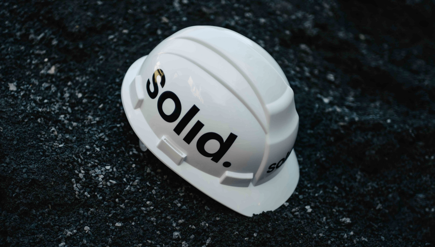 Casque de sécurité blanc avec le logo 'solid.' noir, posé sur un fond texturé sombre, symbolisant la fiabilité et le professionnalisme de la marque.
