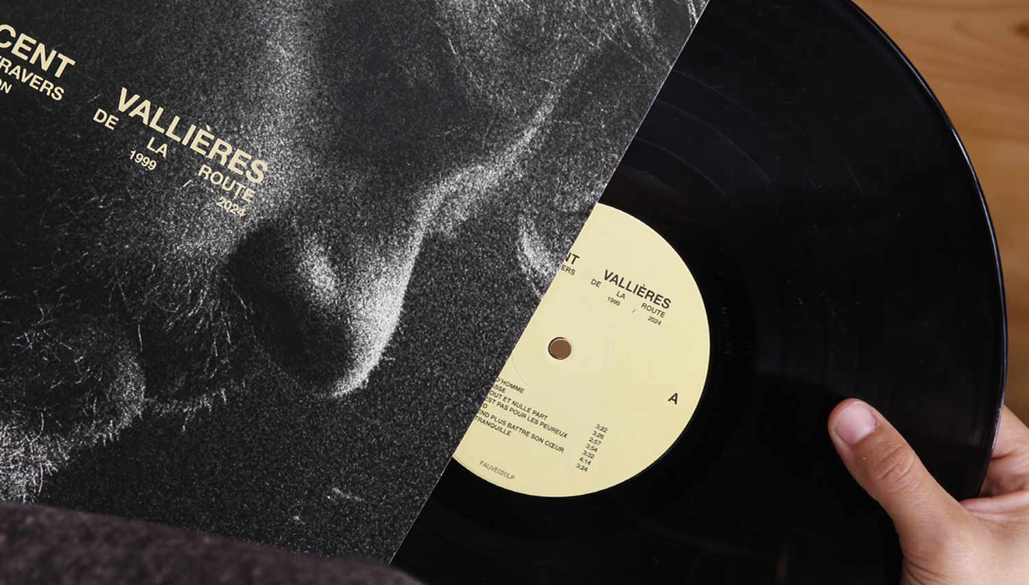 Mise en situation d'un disque vinyle glissant hors de sa pochette. On y voit le contraste entre la pochette cartonnée noir et blanc et le vinyle noir brillant avec son étiquette centrale jaune pâle, tenu par une main.