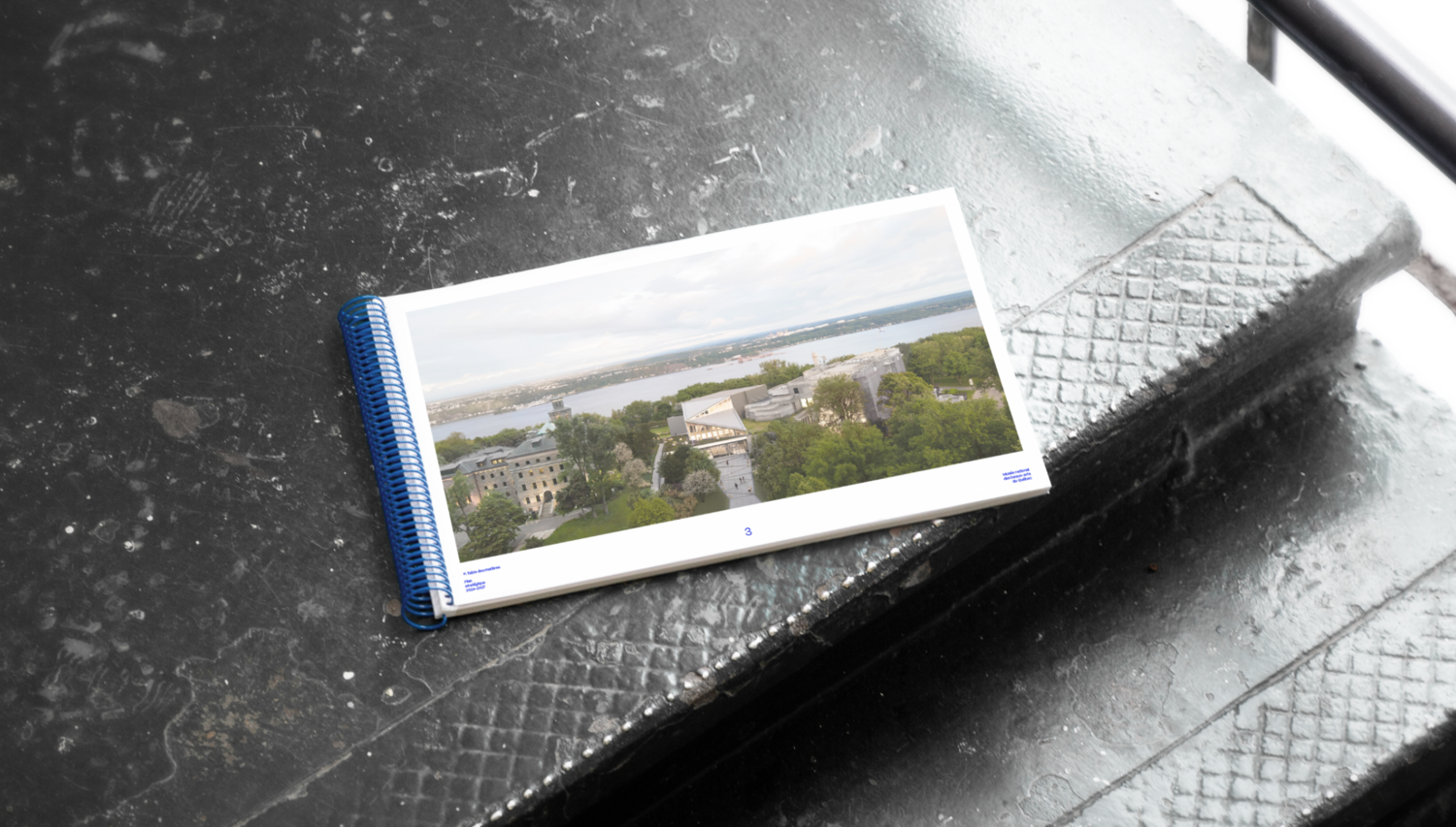 Le document imprimé du plan stratégique ouvert, posé sur une surface texturée sombre, révélant une photographie panoramique du complexe muséal et du fleuve Saint-Laurent.