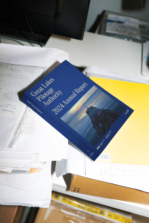 La couverture du rapport annuel 2024 de l’Administration de pilotage des Grands Lacs est posée sur un bureau encombré de documents. Cette mise en scène suggère un environnement de travail actif et l'importance du document.