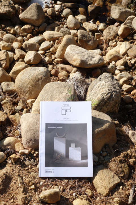 Un exemplaire du magazine Formes est posé sur un lit de galets, sa couverture présentant une œuvre au design épuré. L'image symbolise la rencontre entre l'architecture contemporaine et les matériaux naturels bruts.