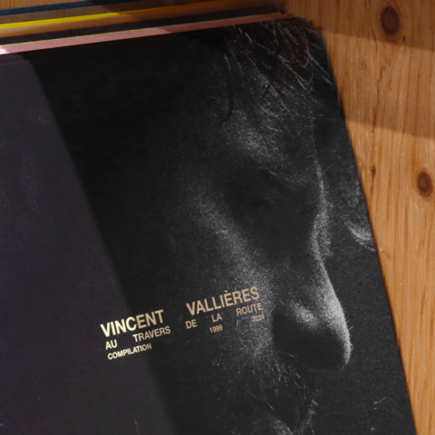 Gros plan artistique sur la couverture de l'album, montrant une photographie très texturée et granuleuse en noir et blanc du profil de Vincent Vallières. Le titre « Au travers de la route » et les années « 1999 / 2024 » apparaissent en typographie dorée contrastante.
