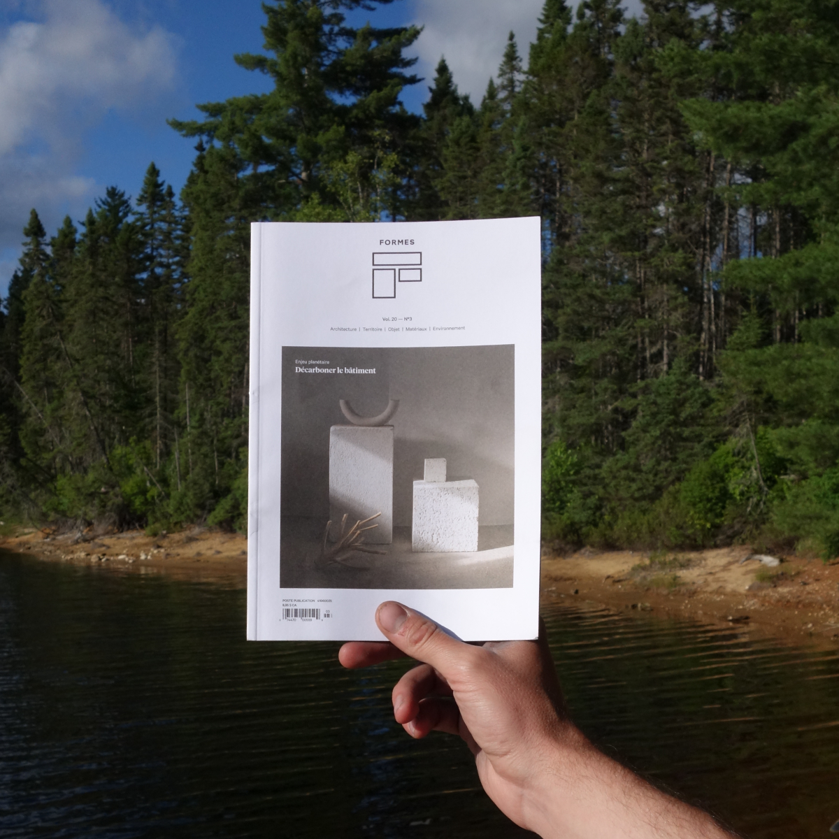 Le magazine Formes est tenu à la main devant un paysage de lac et de forêt typique du Québec. L'image illustre l'inspiration que puise l'architecture écologique dans la nature environnante.