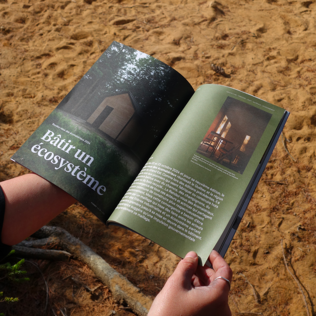 Une personne tient le magazine Formes ouvert sur un fond de sable, lisant l'article « Bâtir un écosystème ». Cette vignette incarne l'invitation à réfléchir sur la construction durable, même en plein air.