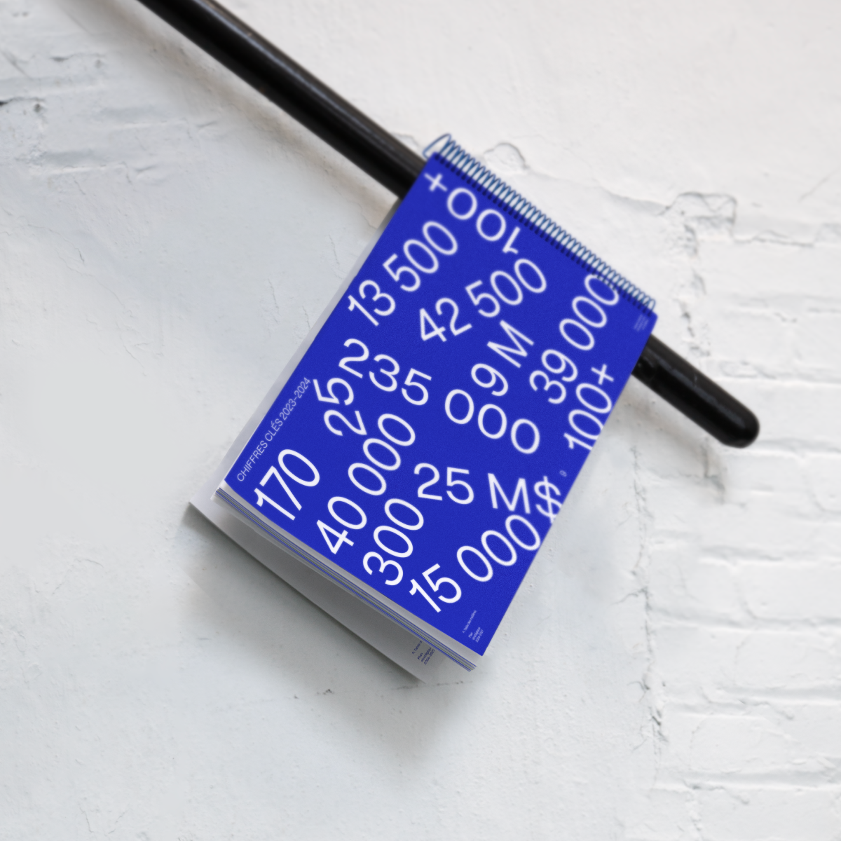 Le document du plan stratégique, ouvert à la page des chiffres clés sur fond bleu, disposé artistiquement sur une rampe noire devant un mur de briques blanches peint.